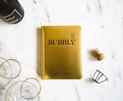 bubbly book with gold cover set on a marble surface with glasses and a bottle of champagne.