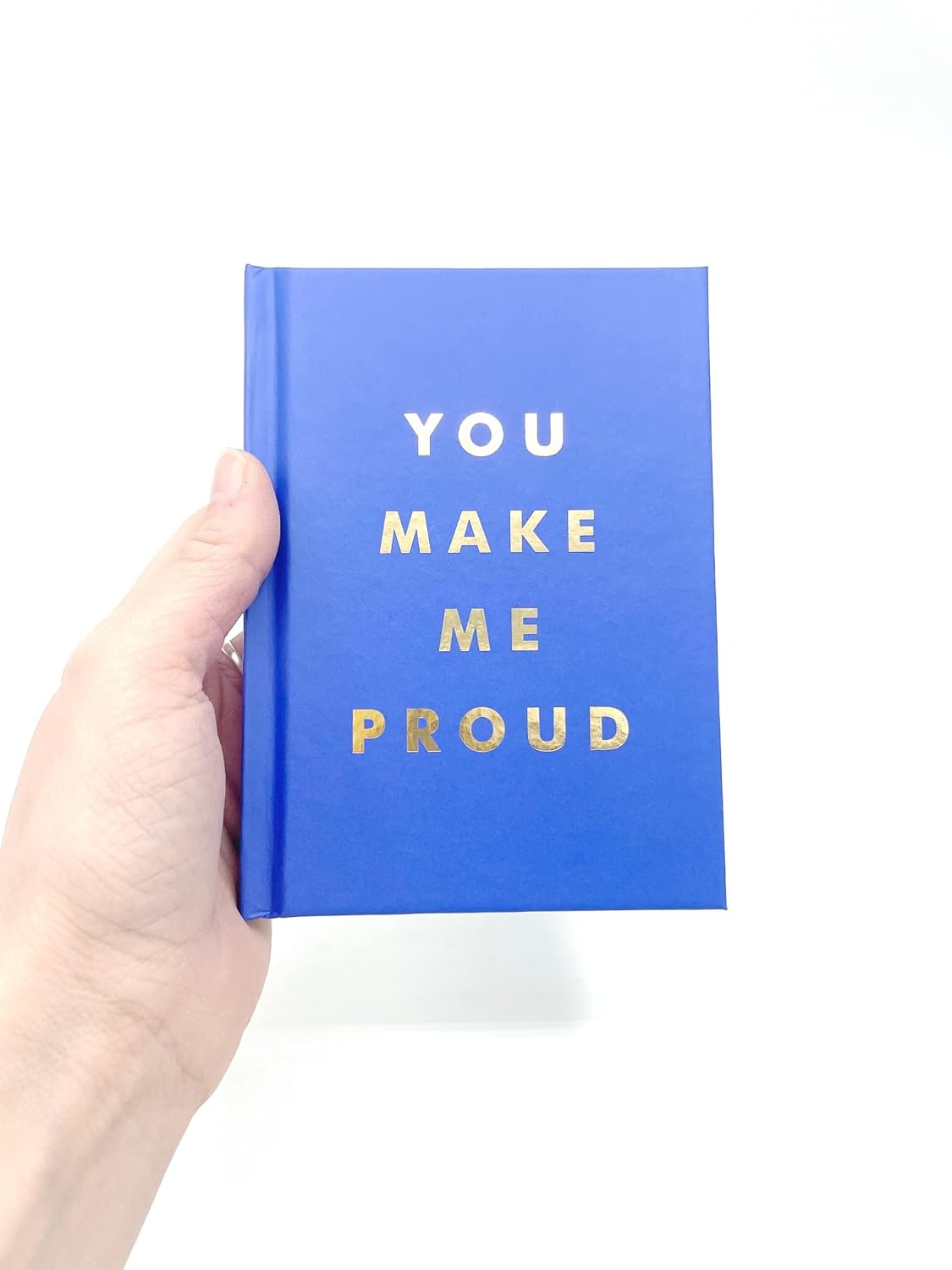 Hand holding a blue book with 'YOU MAKE ME PROUD' text on a white background