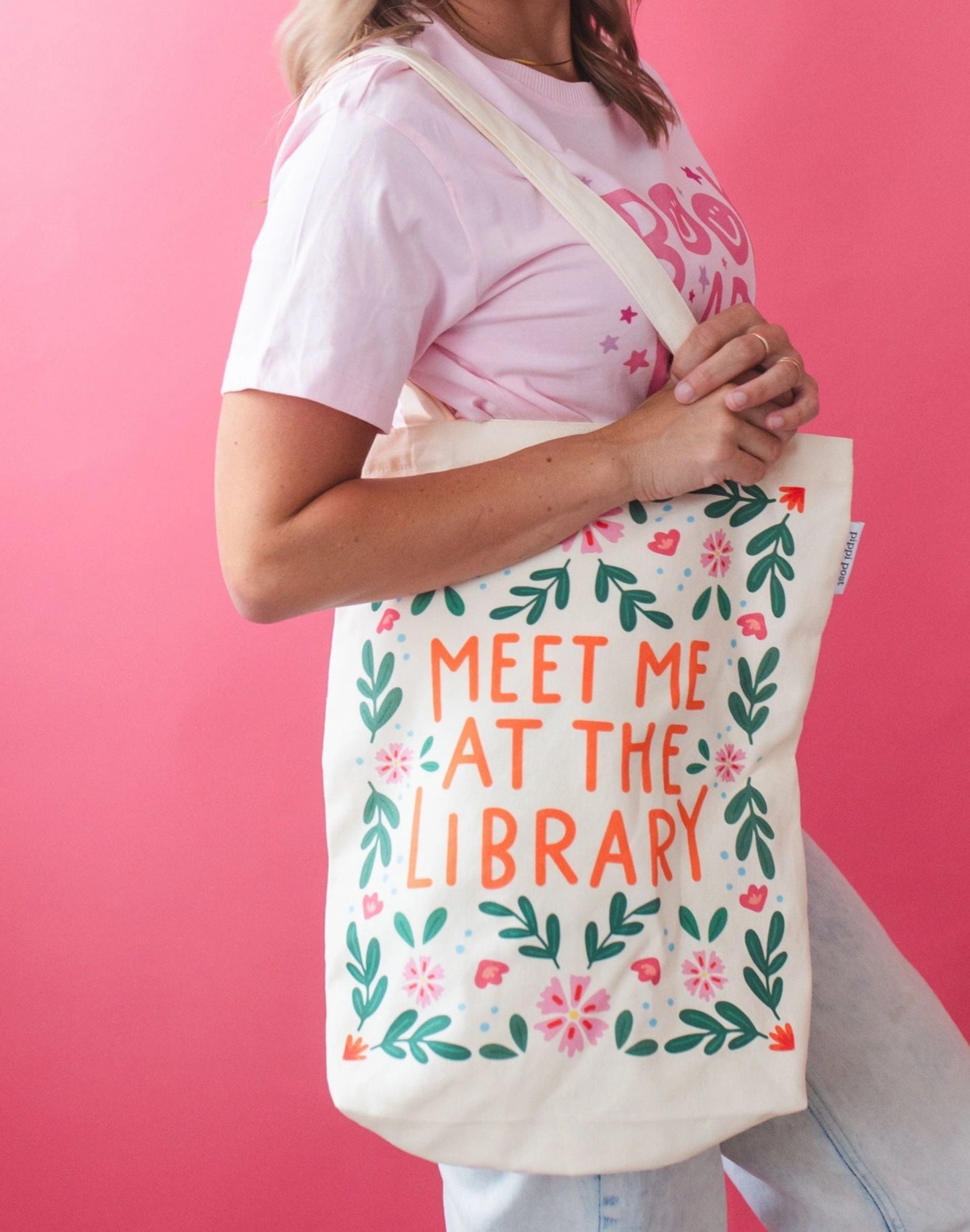 Person holding a tote bag with 'Meet Me at the Library' text against a pink background