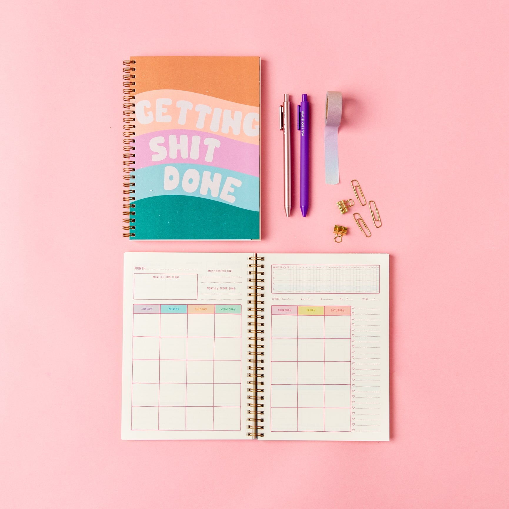 getting shit done spiral planner open and closed on a pink surface with pens, tape, and paperclips.