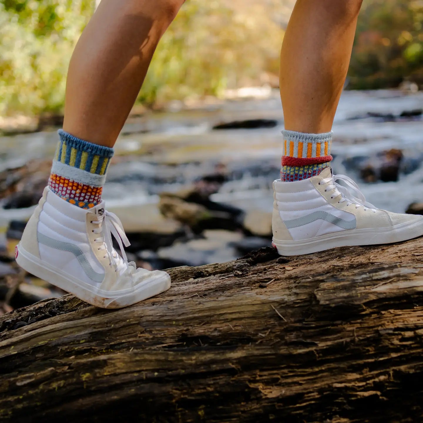 person wearing white high-top sneakers and Fig Crew Socks walking on a log across a river