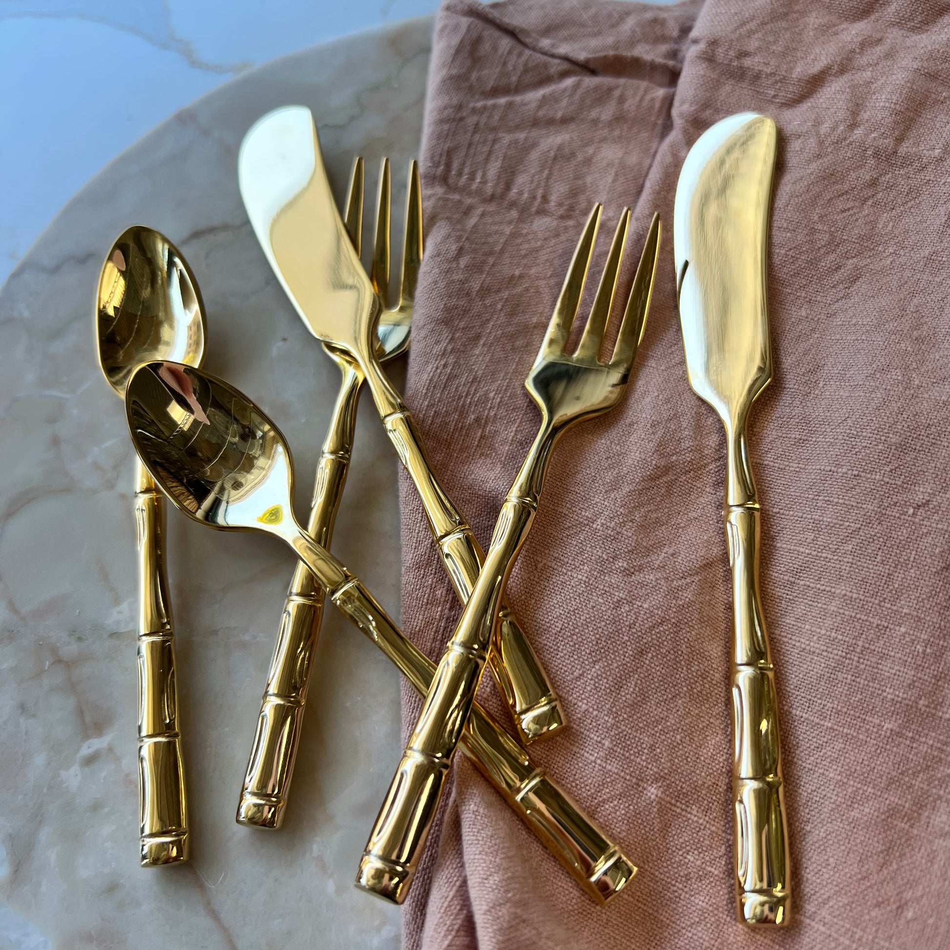 small gold sliver-ware with bamboo-style handles arranged on a marble board with a blush napkins.