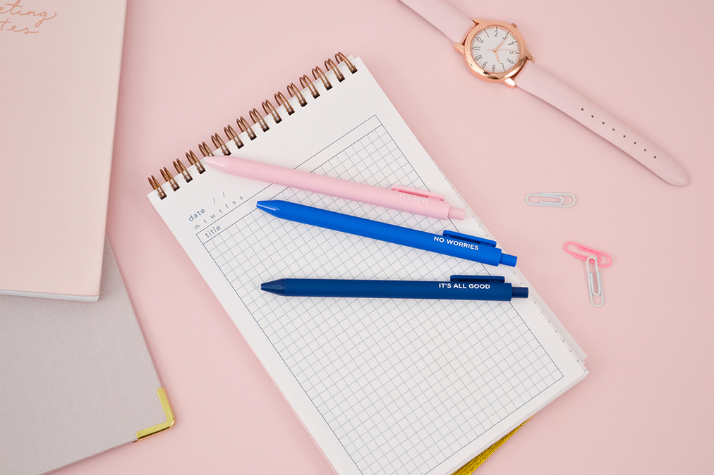 3 all good jotter pens laying on a notebook set on a pink desk with a watch and paperclips.