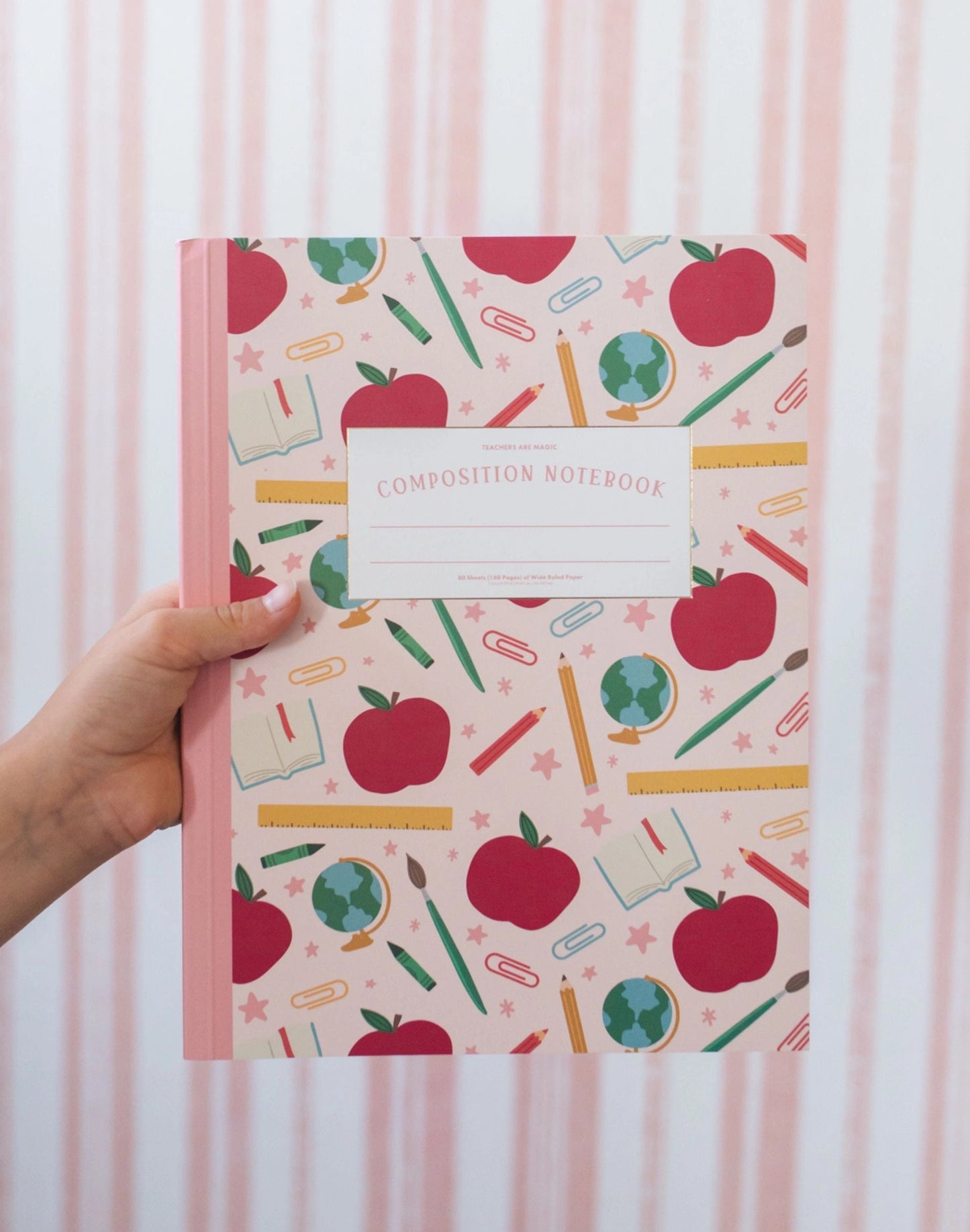 Composition notebook with apple and school supplies pattern held by a hand against a striped background