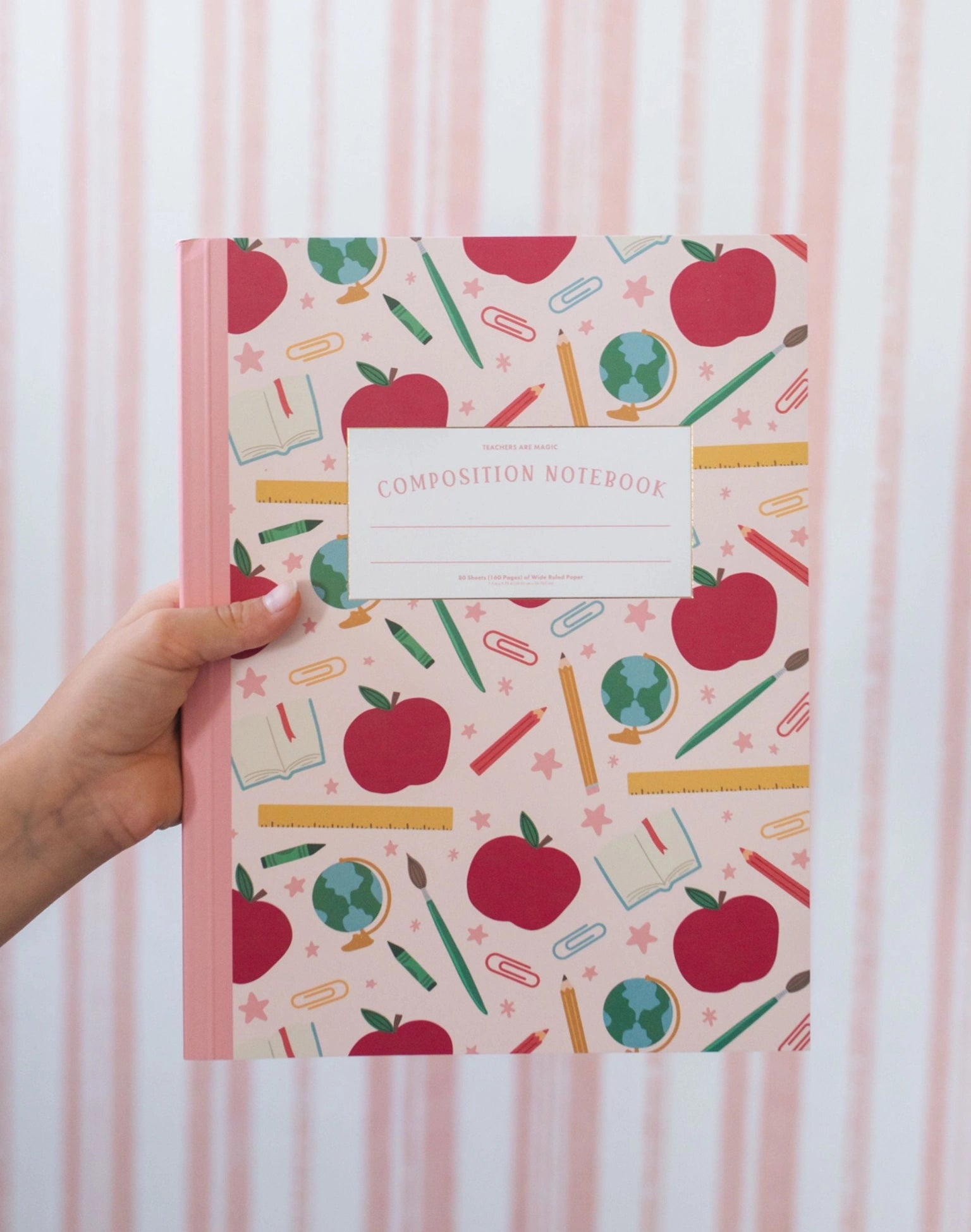 Composition notebook with apple and school supplies pattern held by a hand against a striped background