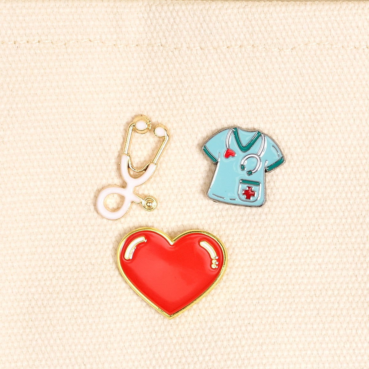 Three pins on a beige fabric background: a stethoscope, a red heart, and a blue medical shirt.