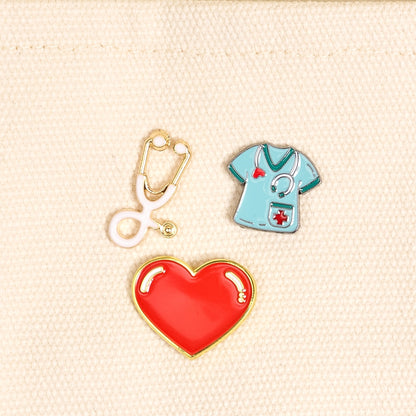 Three pins on a beige fabric background: a stethoscope, a red heart, and a blue medical shirt.