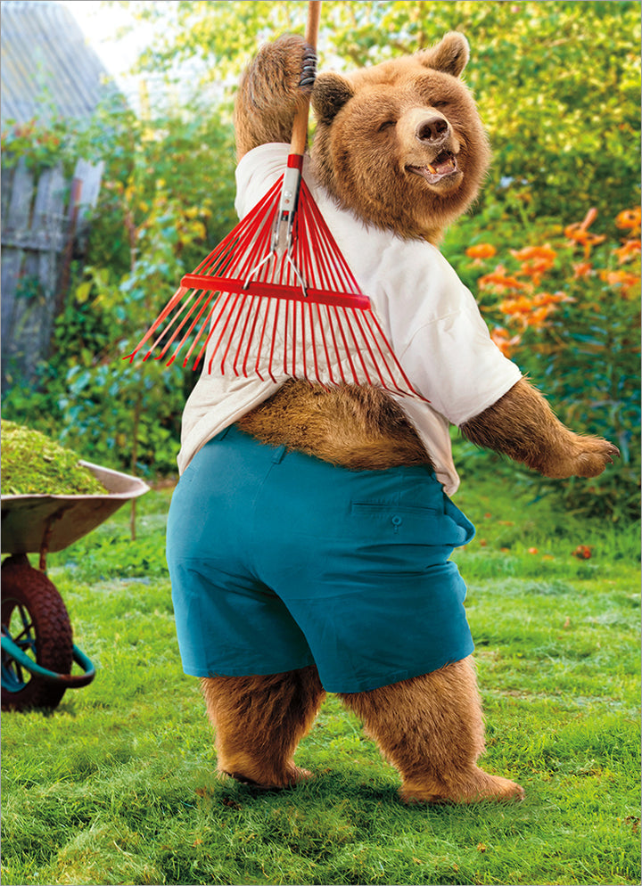 front of card has a bear wearing a white tshirt and blue shorts scratching his back with a red rake while standing outside