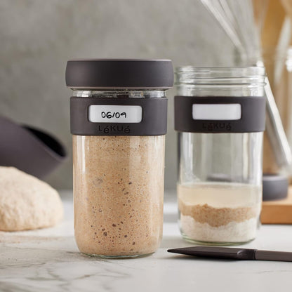 2 jars from the Sourdough Starter Set, one is full and the other partially filled with starter, on a marble counter top.