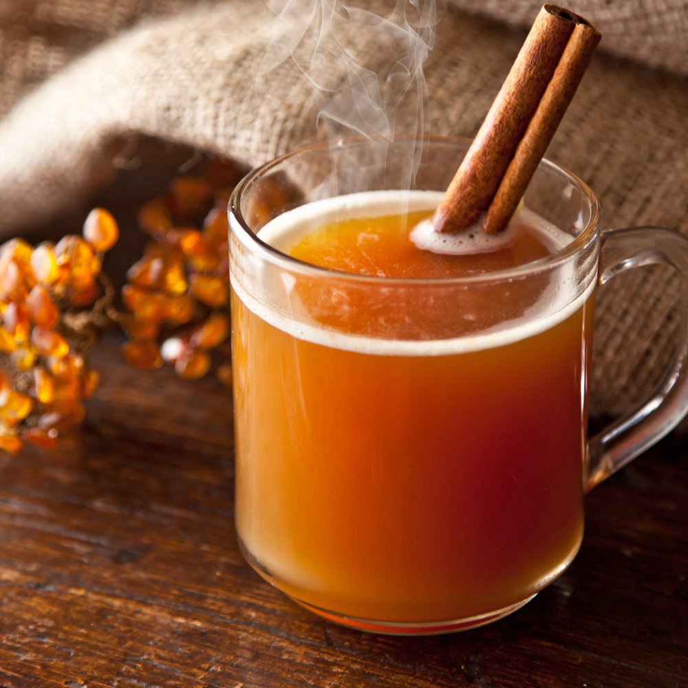Steaming hot beverage in a glass mug with a cinnamon stick on a wooden surface.