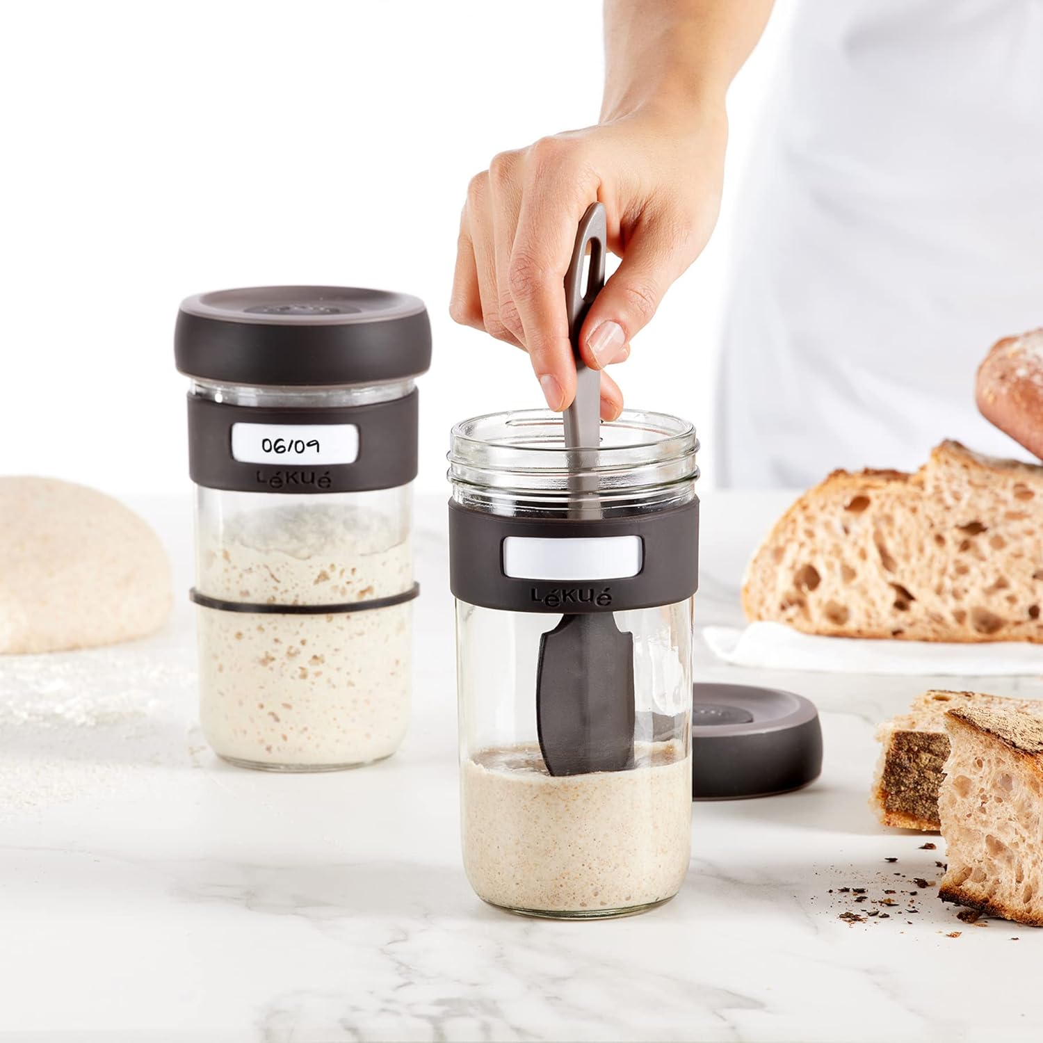 hand using spatula to stir sourdough starter in glass jar set on a counter with bread.