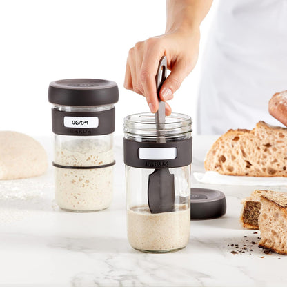 hand using spatula to stir sourdough starter in glass jar set on a counter with bread.