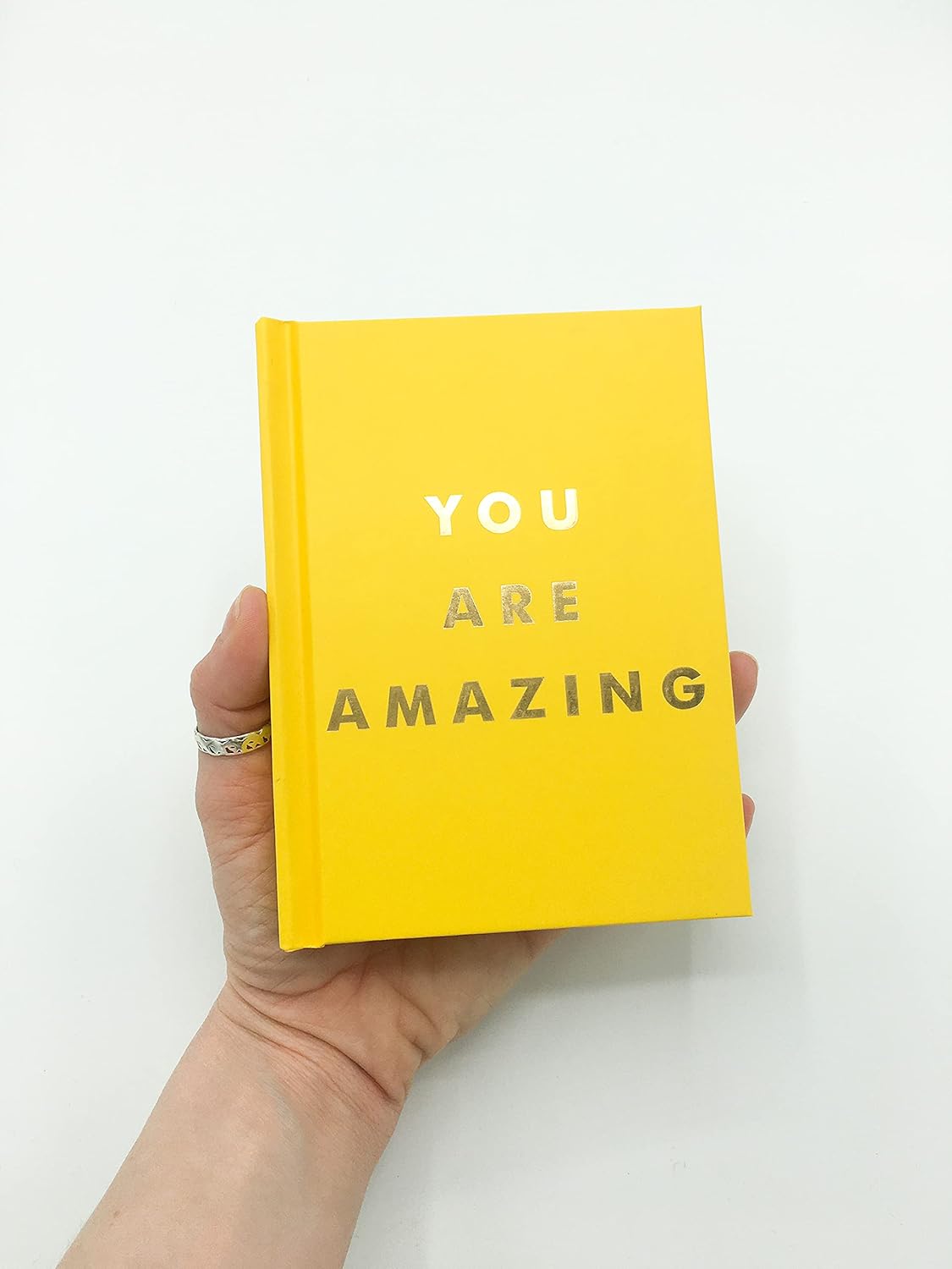 Yellow book with 'YOU ARE AMAZING' text held by a hand on a white background