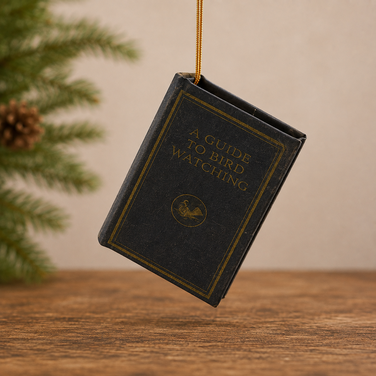 A miniature black book-shaped ornament hanging from a gold string, with the title 'A GUIDE TO BIRD WATCHING' printed in gold on the front, set against a warm beige background with pine branches on the left.