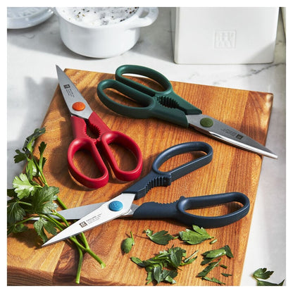 3 kitchen shears laying on a wooden board surrounded by cut fresh herbs.
