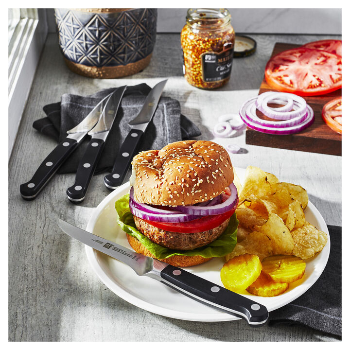plate with a burger, chips, and a PRO Steak Knife on it on a table with other knifes, sliced tomato and onion, and a jar of mustard.