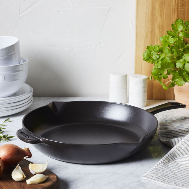 black Cast Iron Fry Pan set on a marble countertop surrounded by a stack of dishes, cutting board with garlic and onions on it. dishtowels, and a pot of greenery.