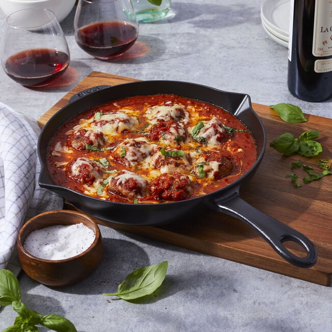 black Cast Iron Fry Pan filled with cooked meatballs set on a wooden board on a table with plates, herbs, and glasses of wine.