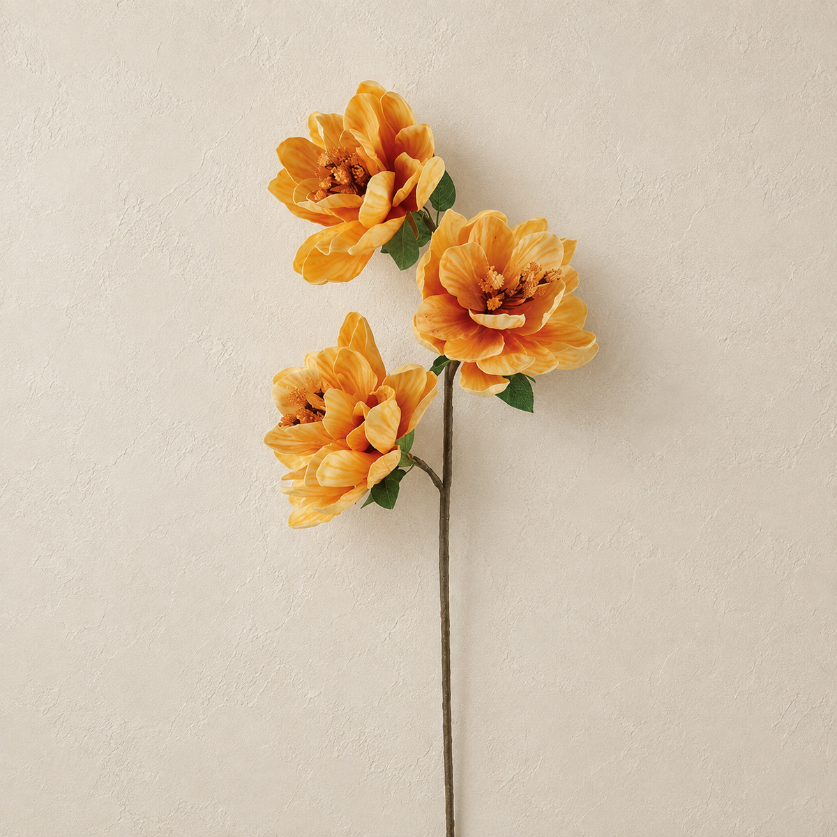 Three artificial orange peony-style flowers with green leaves and stems, arranged against a textured beige wall.