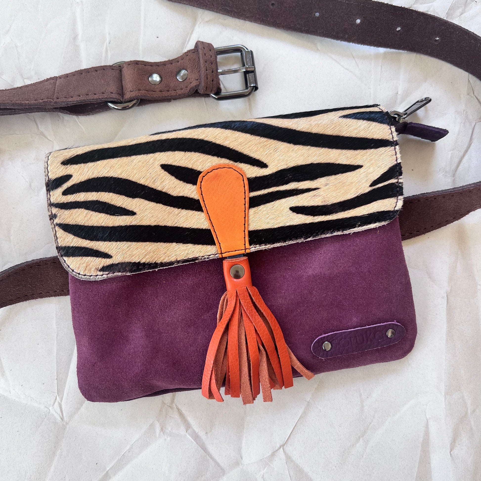 small square bag with purple suede body, black and white stripe animal print flap with orange tassel, and brown belt.