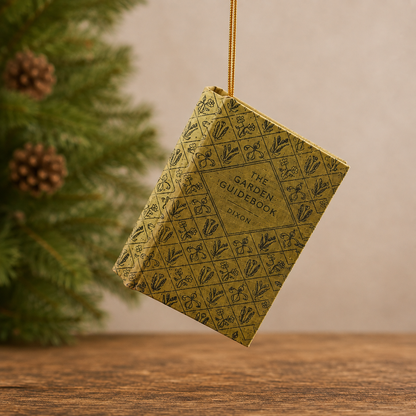 ornament shaped like a vintage hardcover book titled "The Garden Guidebook" by Dixon, suspended by a gold string against a softly blurred indoor background with evergreen branches and a wooden surface.