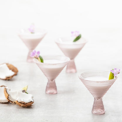 4 pink martini glasses filled with beverage and a floral garnish arranged on a table with bits of coconut.