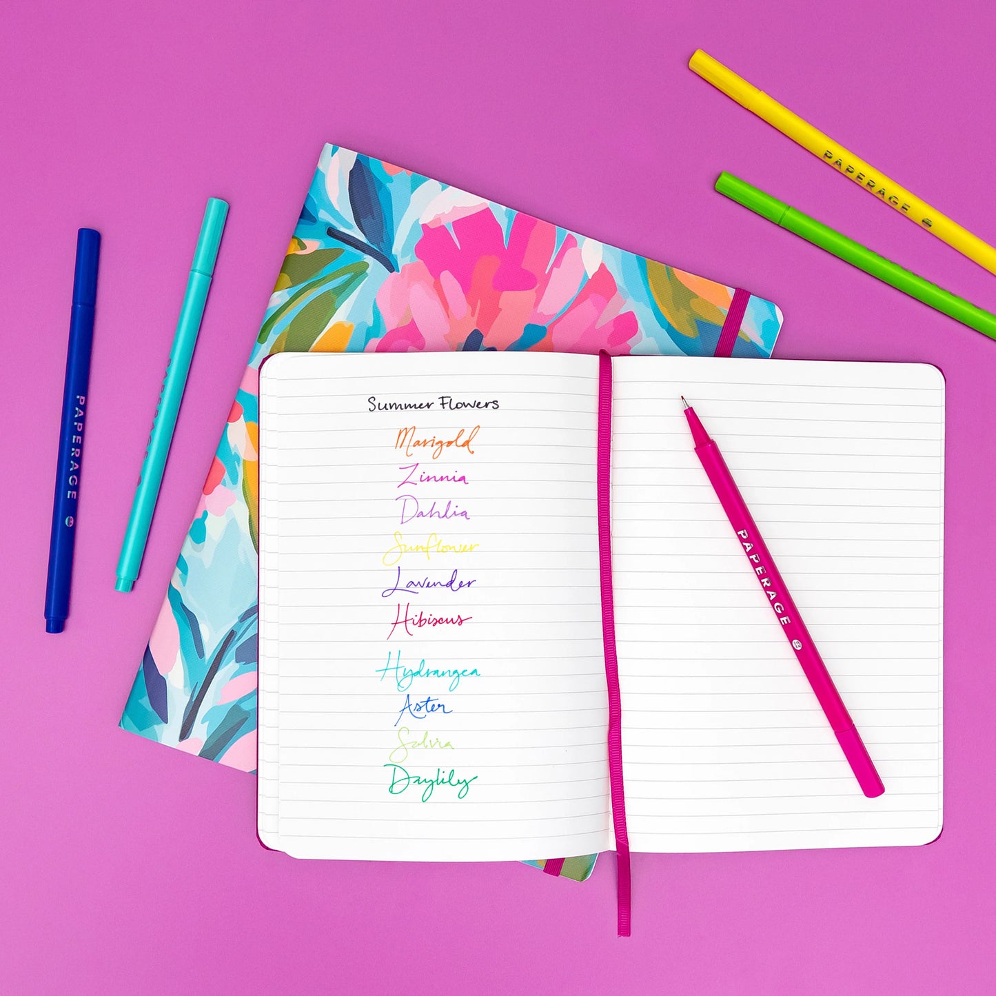 An open lined notebook on a bright pink background, showing colorful handwritten flower names under the heading 'Summer Flowers.' A floral-patterned notebook lies behind it, with two blue pens on the left and colored pencils on the upper right.