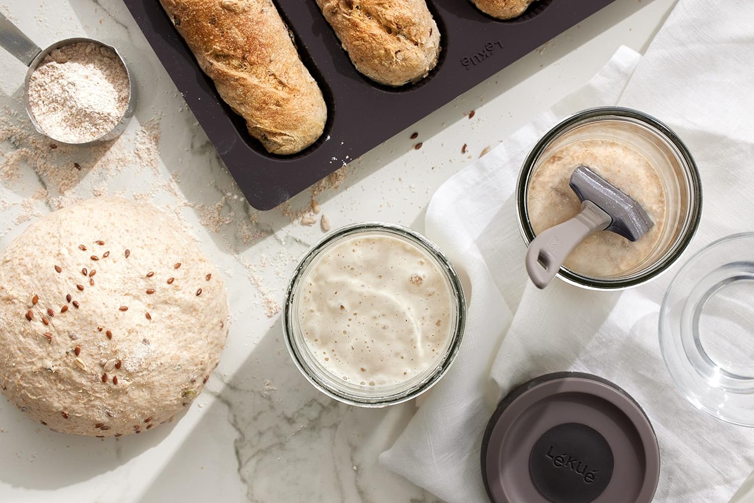 top view of marble countertop arranged with Sourdough Starter Set, pan if baked bread, bread dough, and cup of flour.