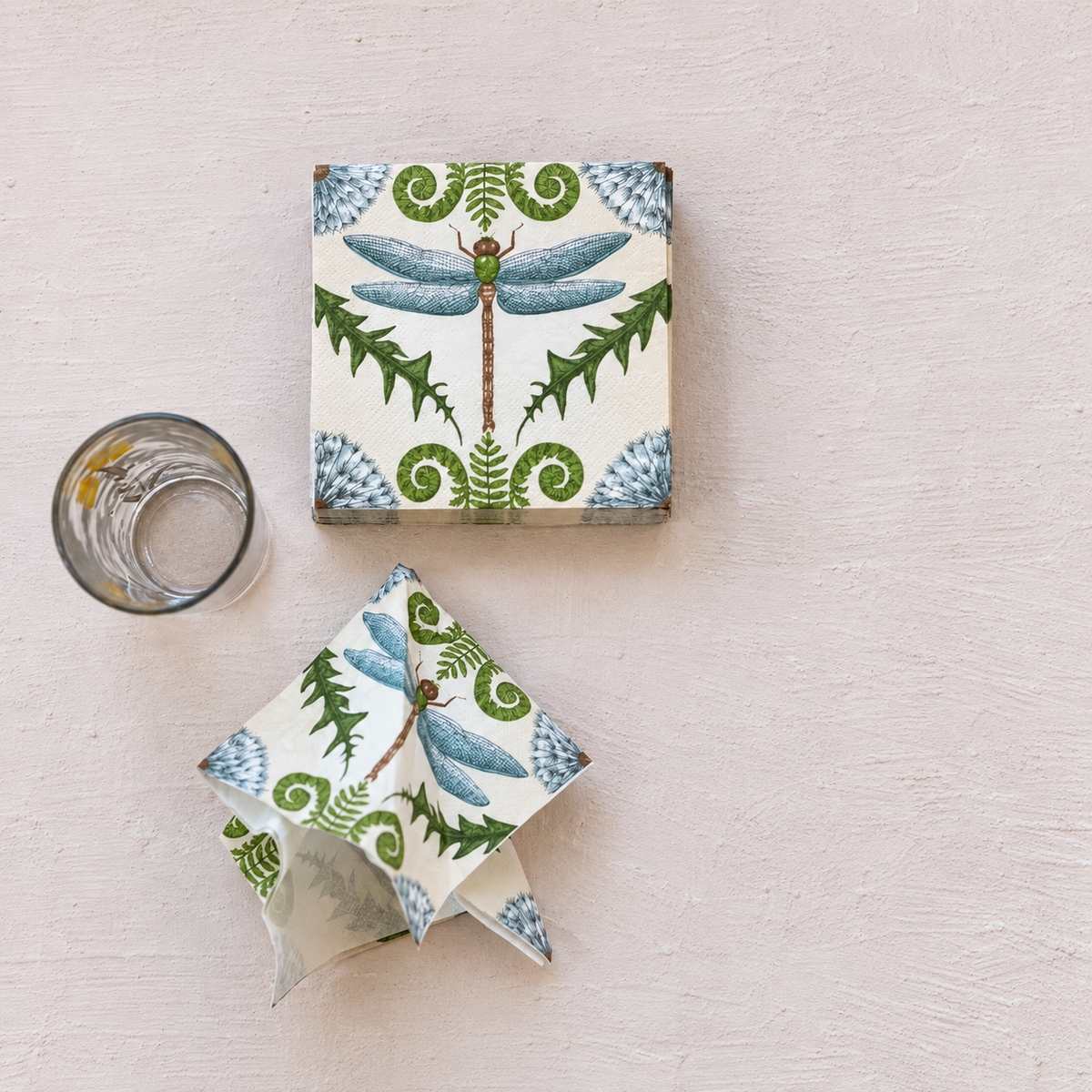 stack of paper napkins with blue and green design of a dragonfly, leaves, and flowers with a crumbled napkin below the stack