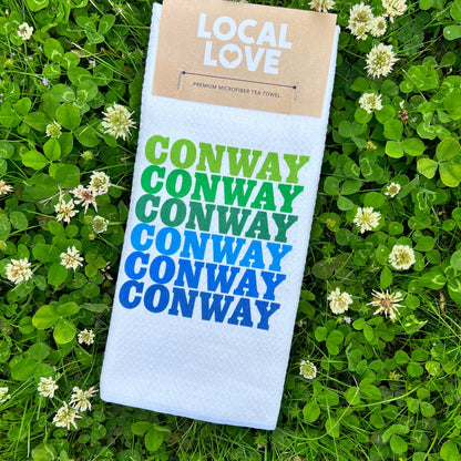 white towel with "conway" printed 6 times in blues and greens laying in a field of clovers.