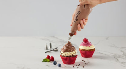 hands holding icing bag and putting chocolate icing on cupcakes set on a marble counter