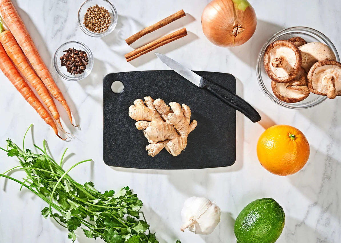 small slate epicurean cutting board with a knife on it on a countertop with fruit, veggies, and spices.