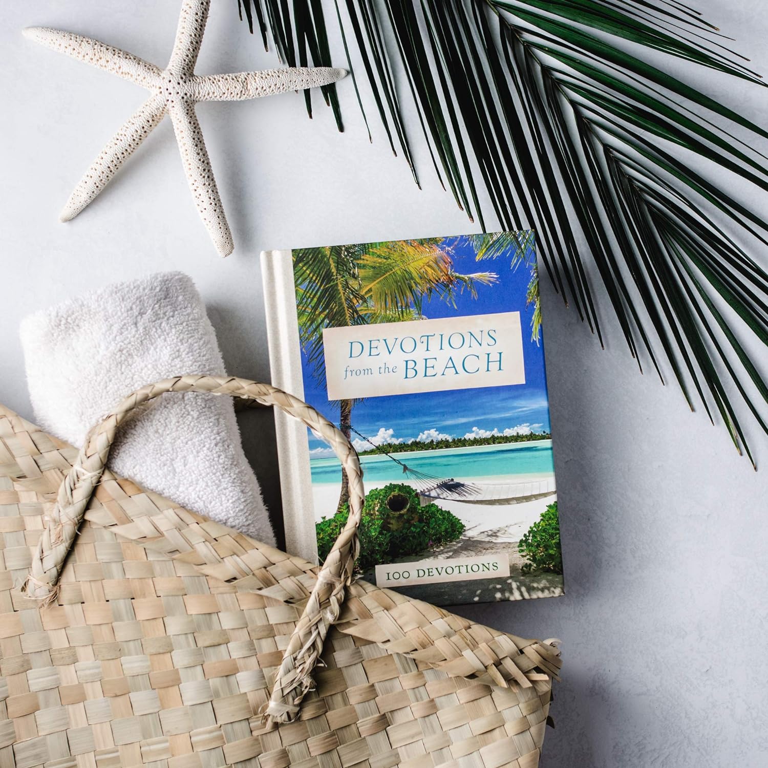 devotions from the beach book laying on a white surface with a beach bag, towel, starfish, and palm leaf.