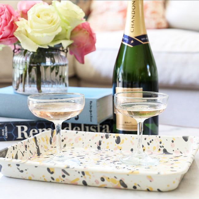 off-white tray with splatters in grey pink and yellow arranged with 2 wine glasses on it and a bottle and books behind it.