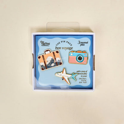 Box of 'bon voyage' themed enamel pins with airplane, suitcase, and camera designs.