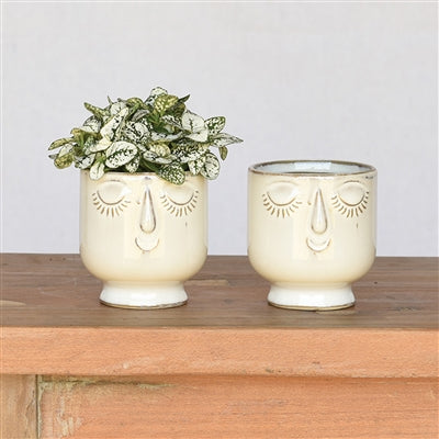 2 ceramic face pots set on a wooden table, one has greenery in it.