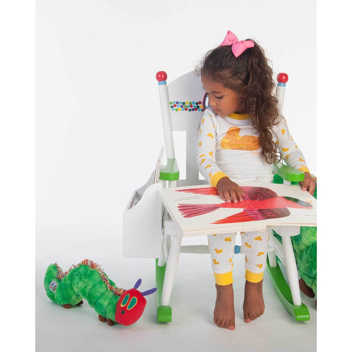 a little girl sitting in a chair holding a book and looking at The Very Hungry Caterpillar Stuffed Animal sitting on the floor