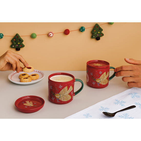 Two red mugs with dove designs on a table, one being picked up, with a plate of cookies and festive background.
