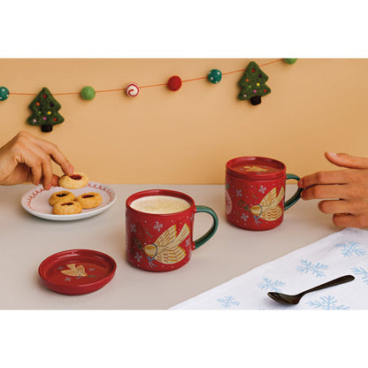 Two red mugs with dove designs on a table, one being picked up, with a plate of cookies and festive background.