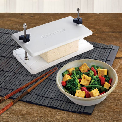 Tofu press with a block of tofu on a wooden table next to a bowl of salad.