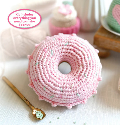 A crocheted pink donut with small pastel sprinkle details, shown on a light fabric surface with a gold spoon, yarn balls, and a blurred cupcake in the background.