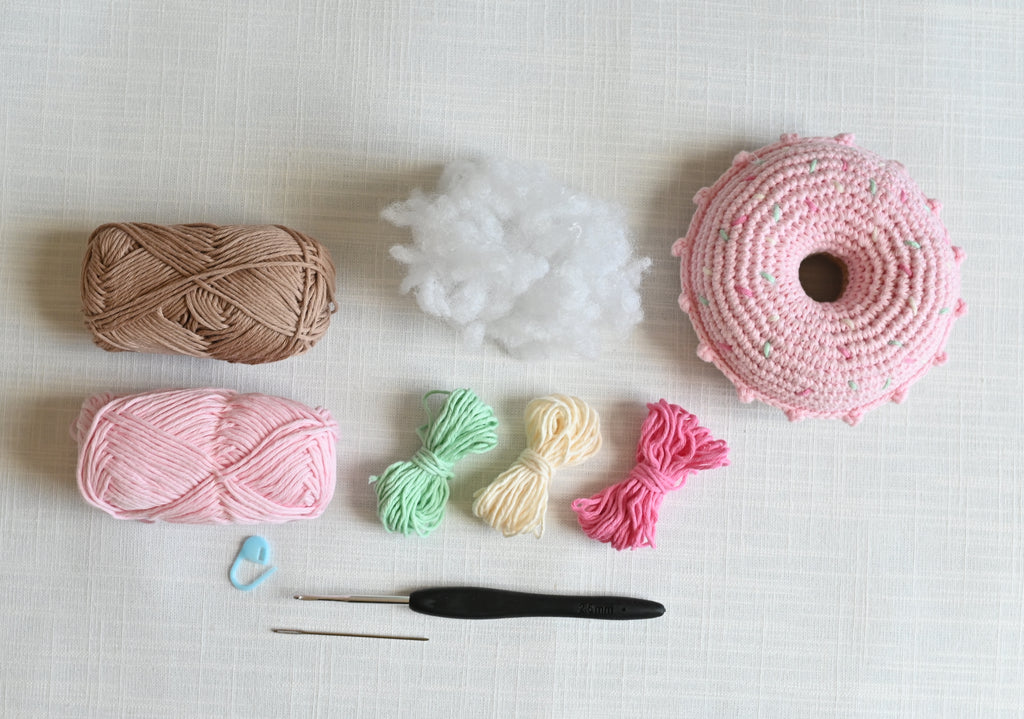 Crocheted donut with a pink and green dots, displayed with its crafting materials including brown and pink yarn, white stuffing, small yarn bundles, a black crochet hook