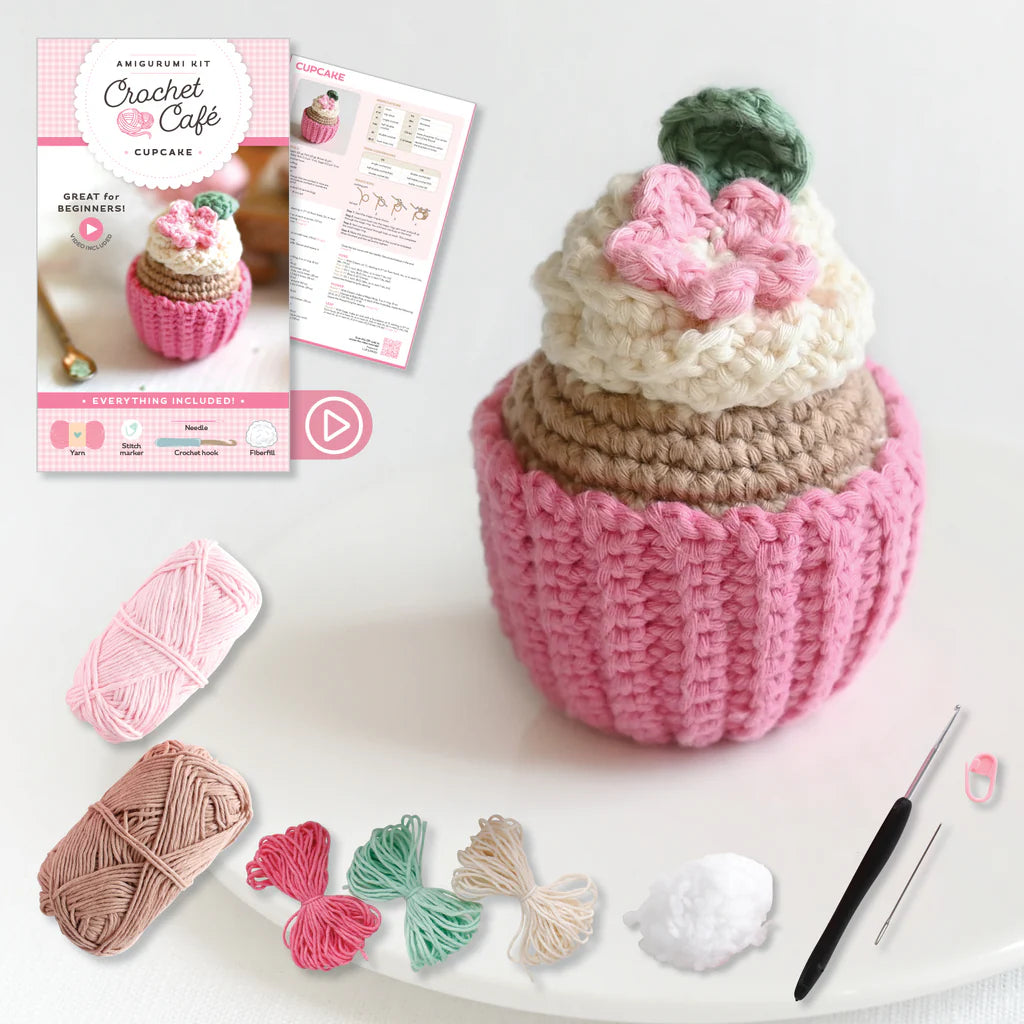 Crocheted cupcake with pink flower, yarn balls, and crochet hook on a white background