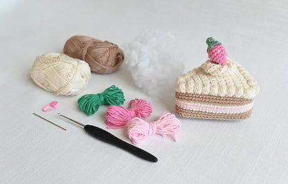Crocheted cake slice craft kit with cream, beige, pink, and green yarn, a black crochet hook, a pink stitch marker, and a white stuffing ball on a white background.