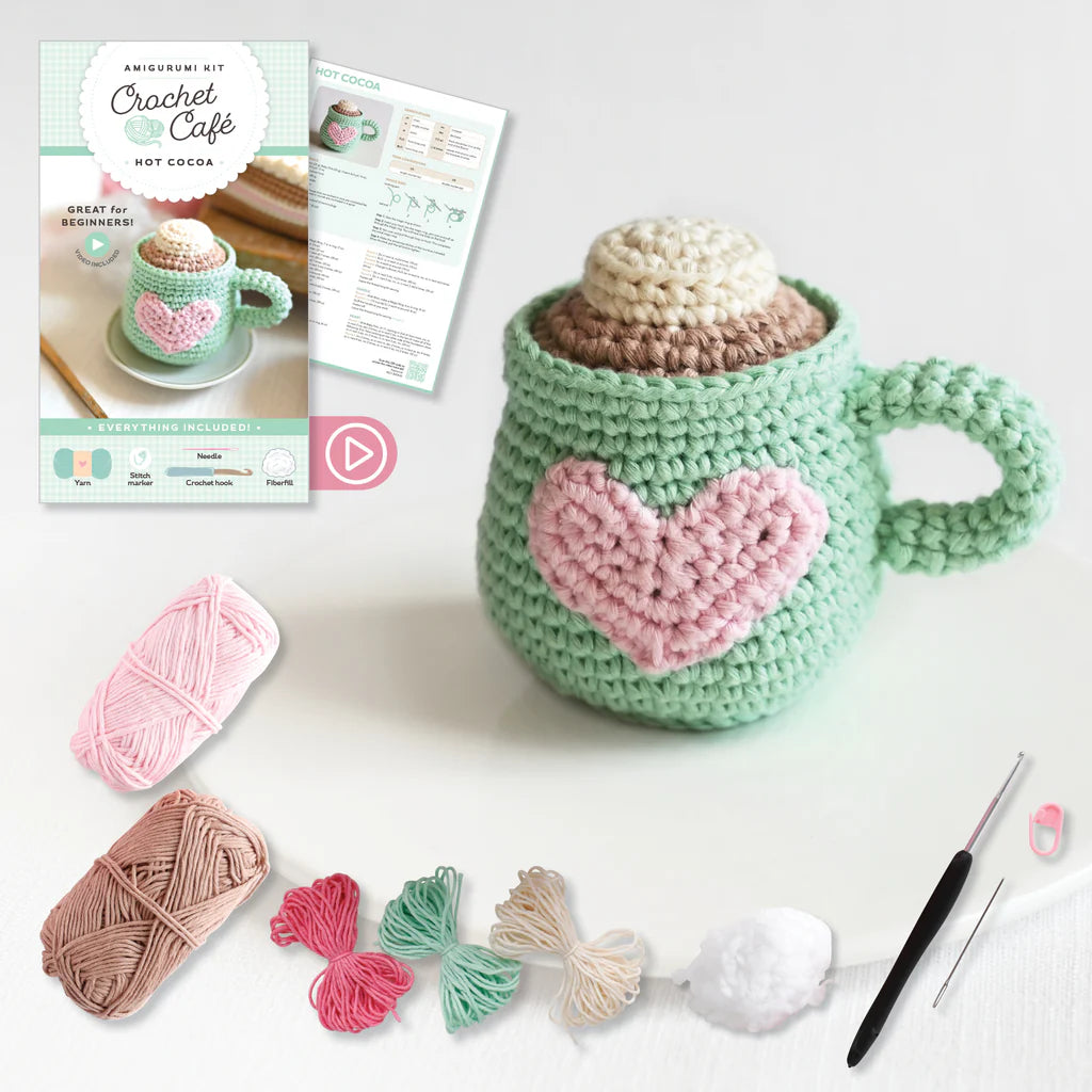 Crocheted mug with heart design, yarn, crochet hook, and pattern book on a white background