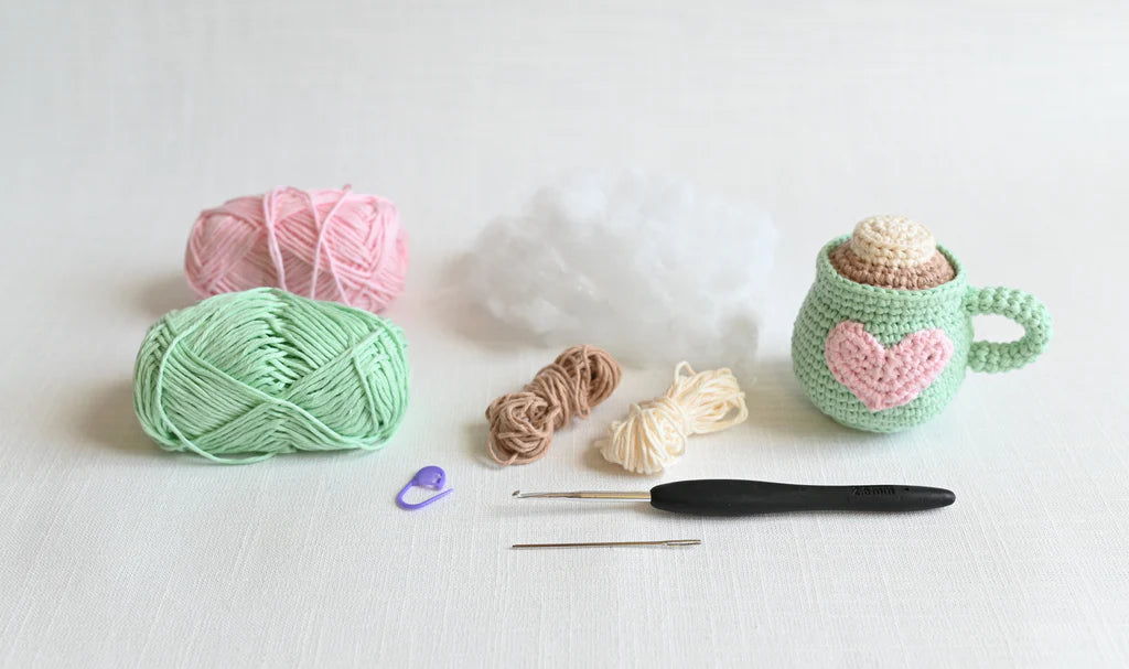 Crochet supplies including yarn, a hook, and a small crocheted green  mug on a white background