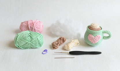 Crochet supplies including yarn, a hook, and a small crocheted green  mug on a white background