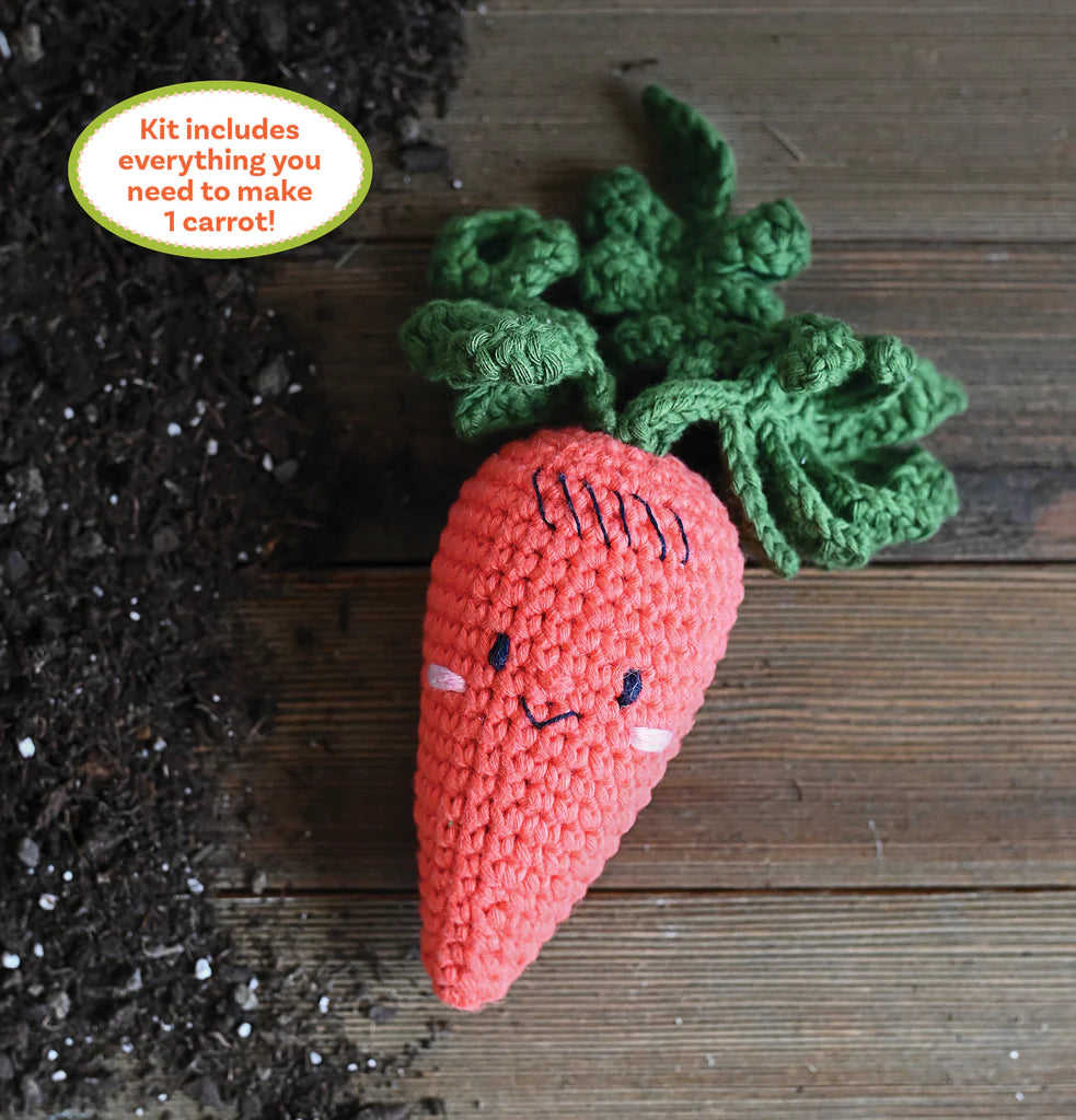 Crocheted carrot with green leaves on a wooden surface next to soil.