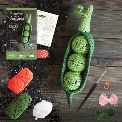 Crocheted green peas in a pod with yarn, needles, and packaging on a wooden surface.
