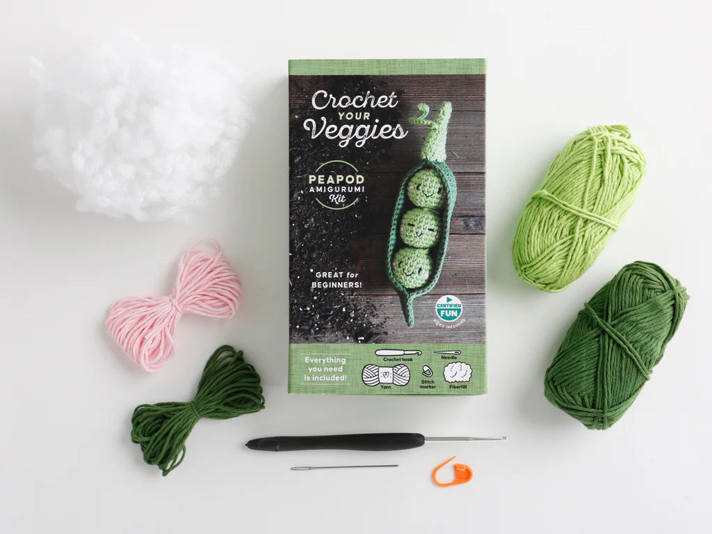  'Crochet Your Veggies' knitting kit on a white background, surrounded by its components: green and pink yarn, a black crochet hook, an orange stitch marker, and a white ball of stuffing. 